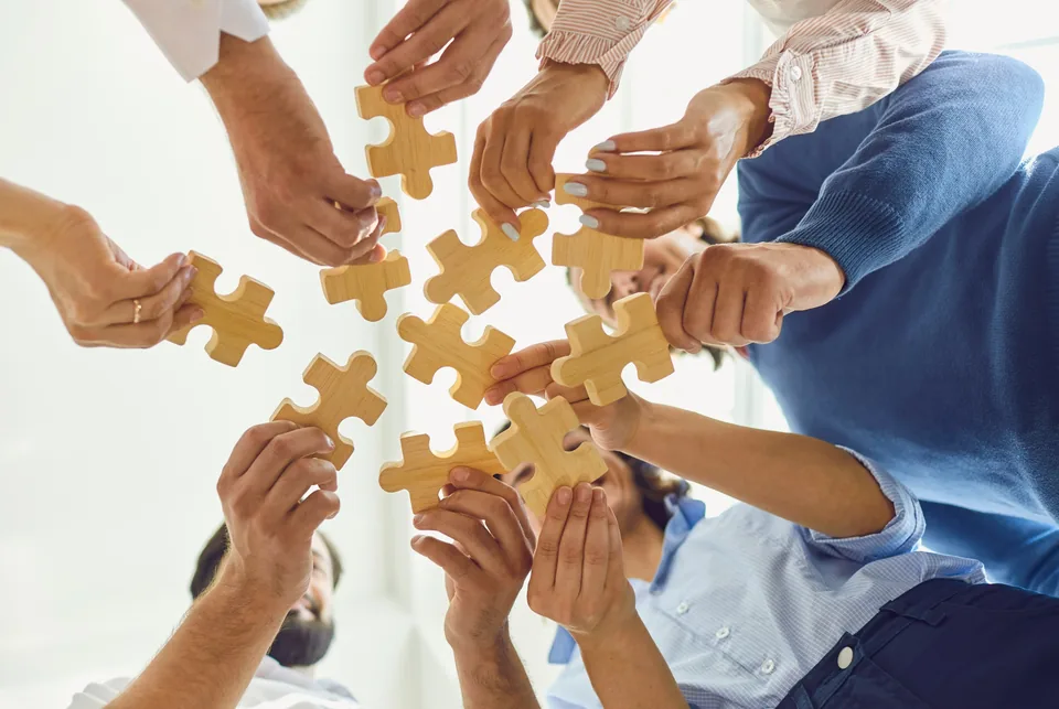 Several people each hold a jigsaw piece, bringing them together towards the centre of a circle.